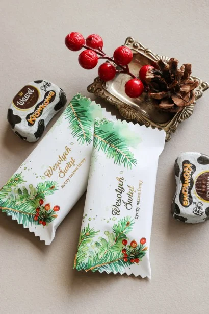 Personalizowane czekoladki świąteczne z logo firmy w eleganckim białym opakowaniu z zielonymi gałązkami i czerwonymi jagodami.