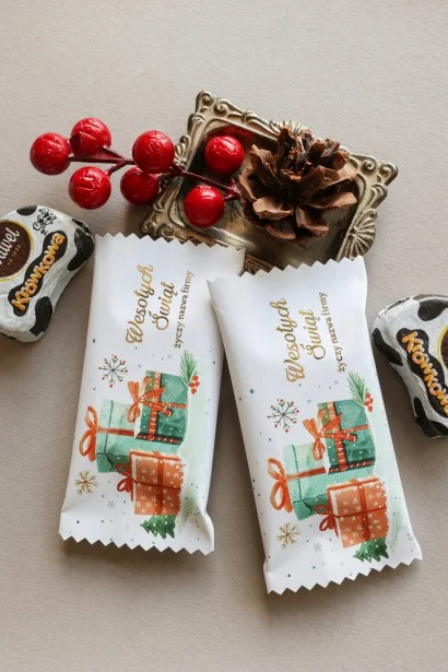 Świąteczne czekoladki w białym opakowaniu z grafiką prezentów, ozdobione zielonymi, czerwonymi i złotymi akcentami.